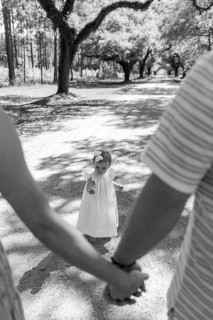 Black and white photo of toddler girl in white dress looking up at parents holding hands outdoors.