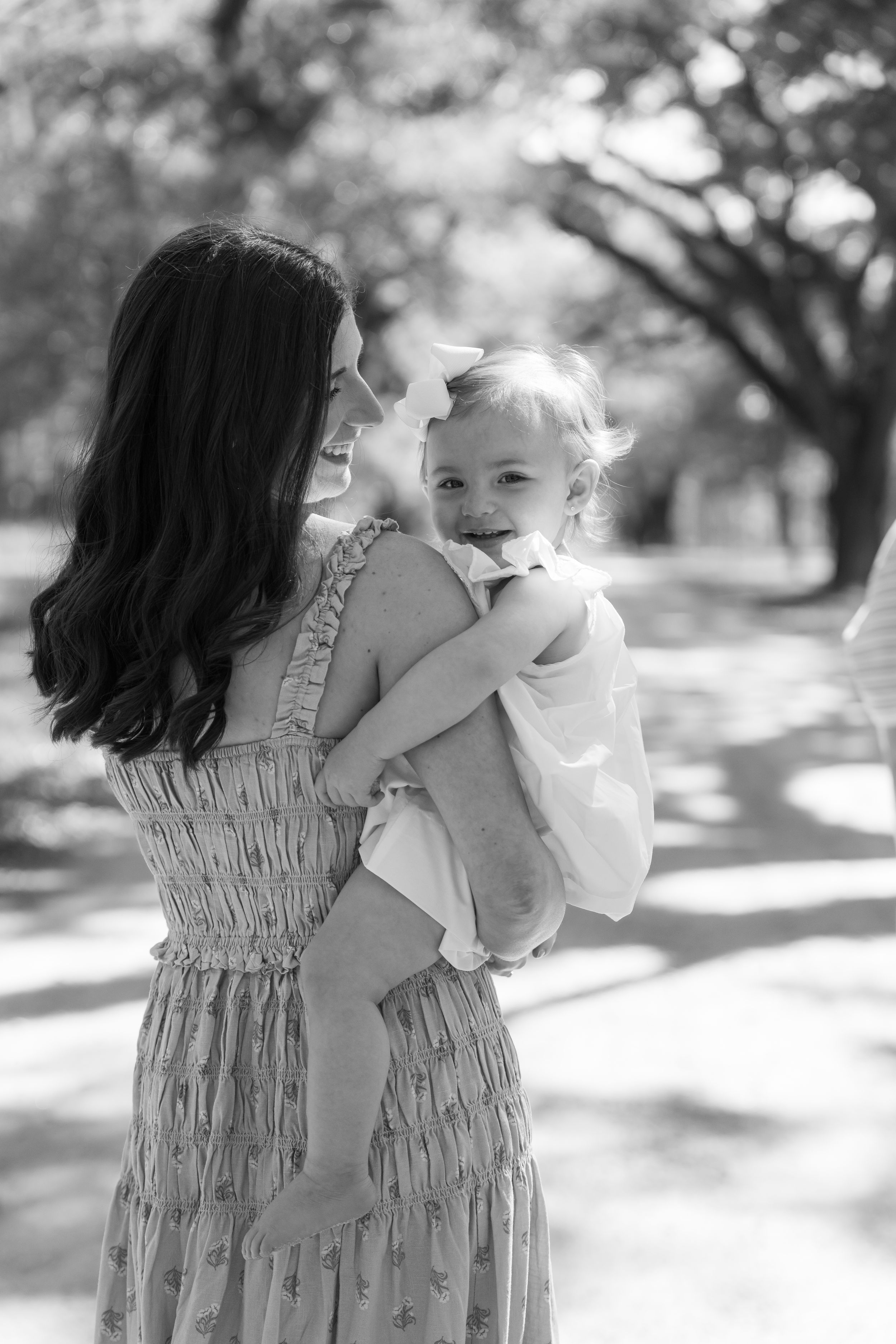 Black and white photo of a smiling baby being held by a dark-haired woman in a floral dress outdoors.