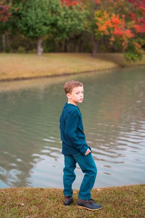 Colorful autumn scene with a small figure in blue walking along a pond with red foliage in background.