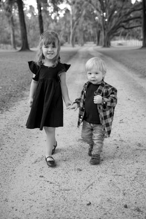 Two small children walk together on a tree-lined path in black and white.