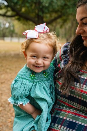 A smiling child wearing a turquoise smocked dress and red hair bow is held by someone in a plaid shirt outdoors.