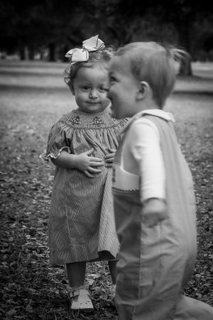 Two toddlers in vintage-style clothing share a sweet moment together on fallen leaves in black and white photography.