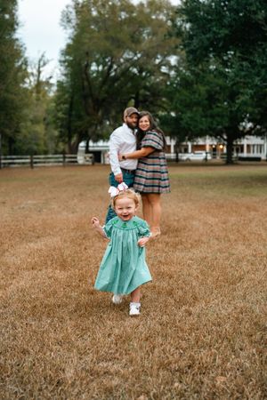 A toddler in a teal dress runs joyfully through a grassy field while a couple embraces in the background.