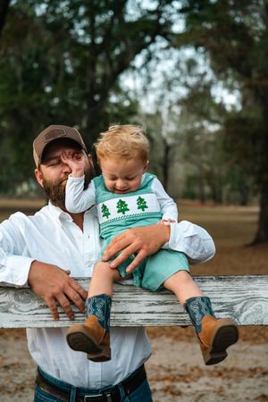 A toddler in a festive green sweater and boots sits on a wooden fence with an adult in casual attire.