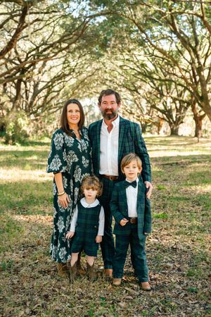 Family in coordinated green plaid and floral outfits poses together under oak trees in a natural outdoor setting.