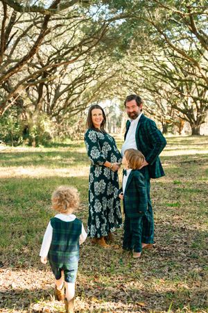 A family in coordinating outfits walks together through a sunlit grove of oak trees with fallen autumn leaves.