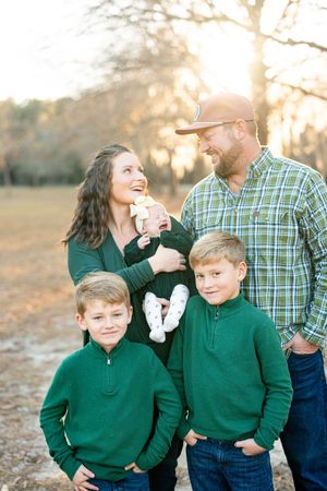 Family wearing matching green sweaters and jeans poses together outdoors at sunset in a field.