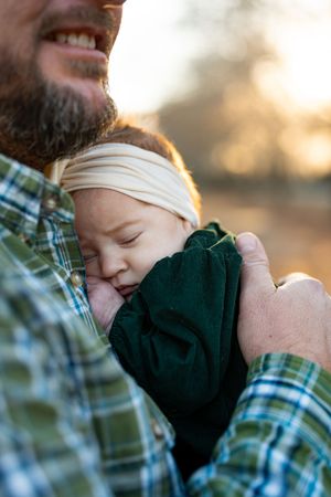 A sleeping baby in a dark green outfit is cradled in plaid-shirted arms during golden hour sunset.