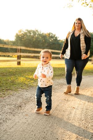 A toddler walks along a path during golden hour wearing jeans and a patterned shirt.