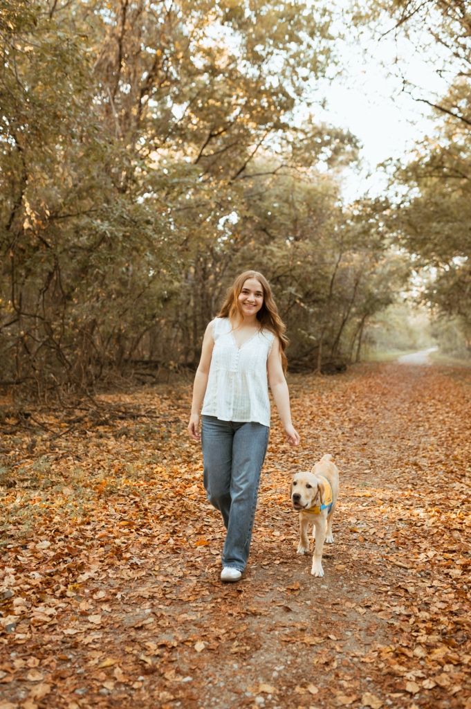 Person walking with a dog through autumn leaves on a wooded path.