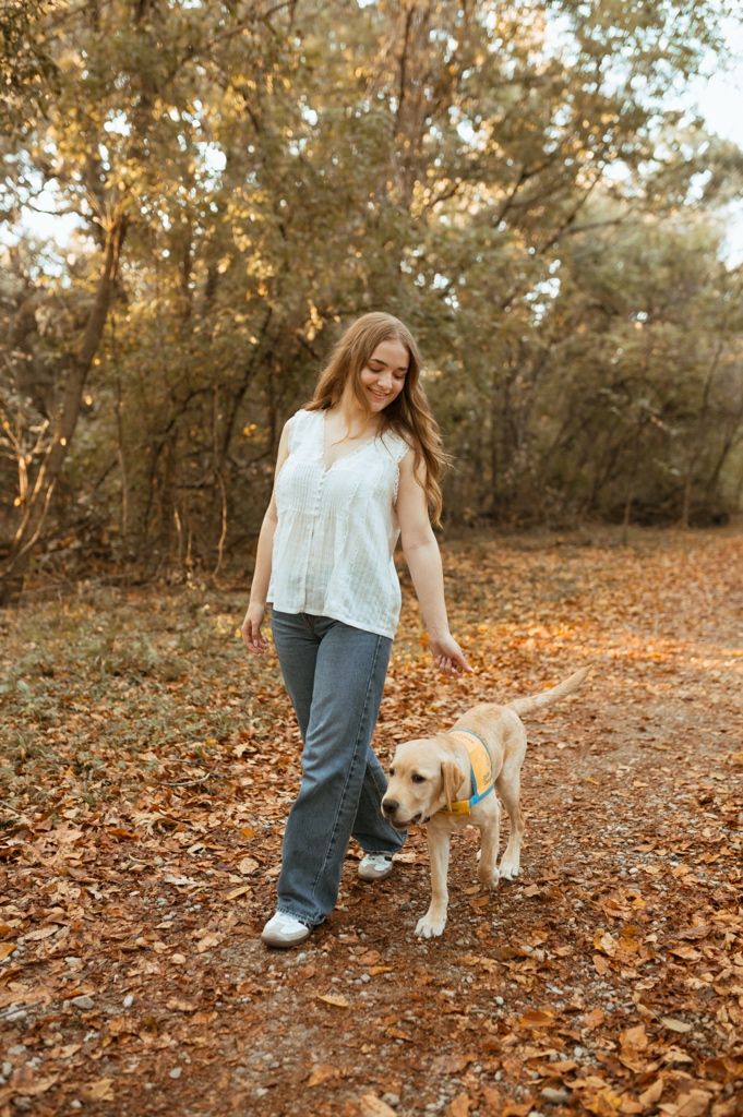 A person in jeans and a light top walking with their yellow lab puppy through autumn foliage.