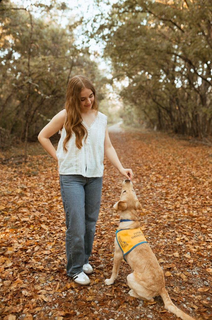 A service dog in a yellow vest interacts with their companion while standing on a leaf-covered forest trail.