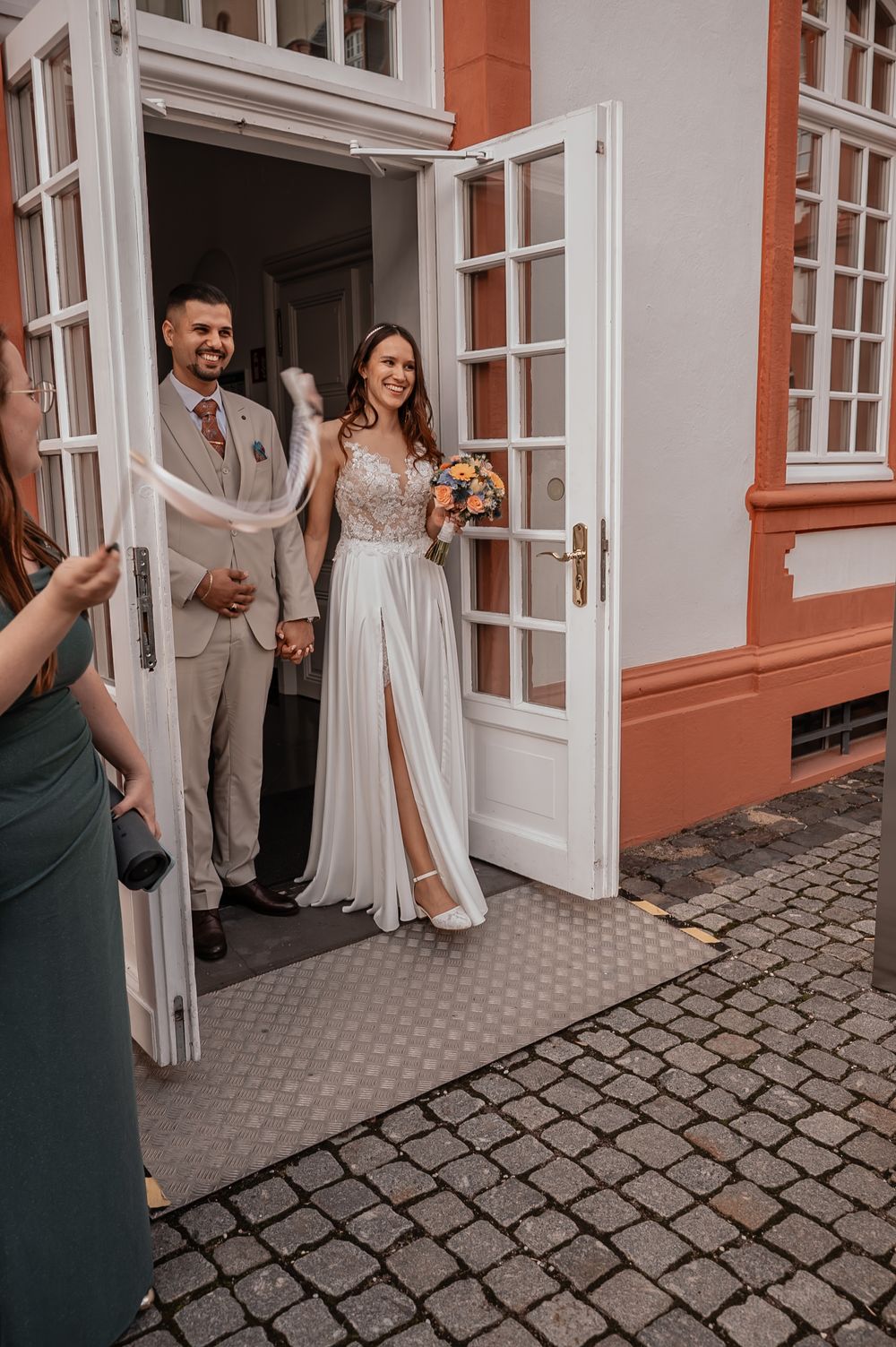 A wedding couple poses in front of a historic building with white doors and cobblestone street.