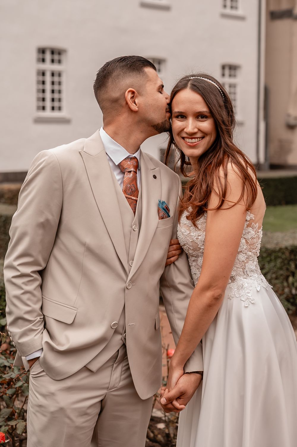 Newlyweds in white dress and beige suit share intimate moments during their outdoor wedding photoshoot.