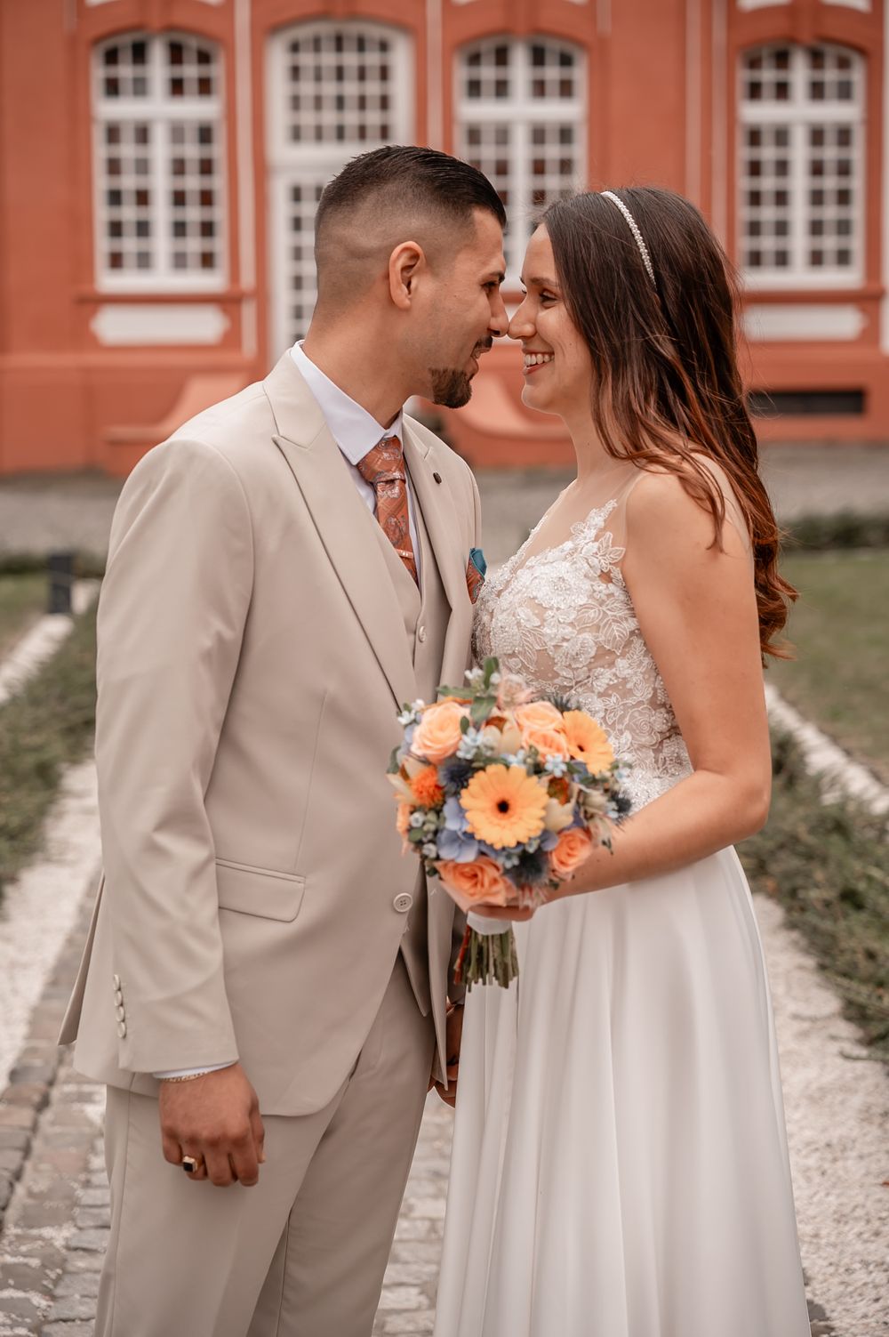 Romantic wedding couple in beige suit and white dress share intimate moments against a brick building backdrop.
