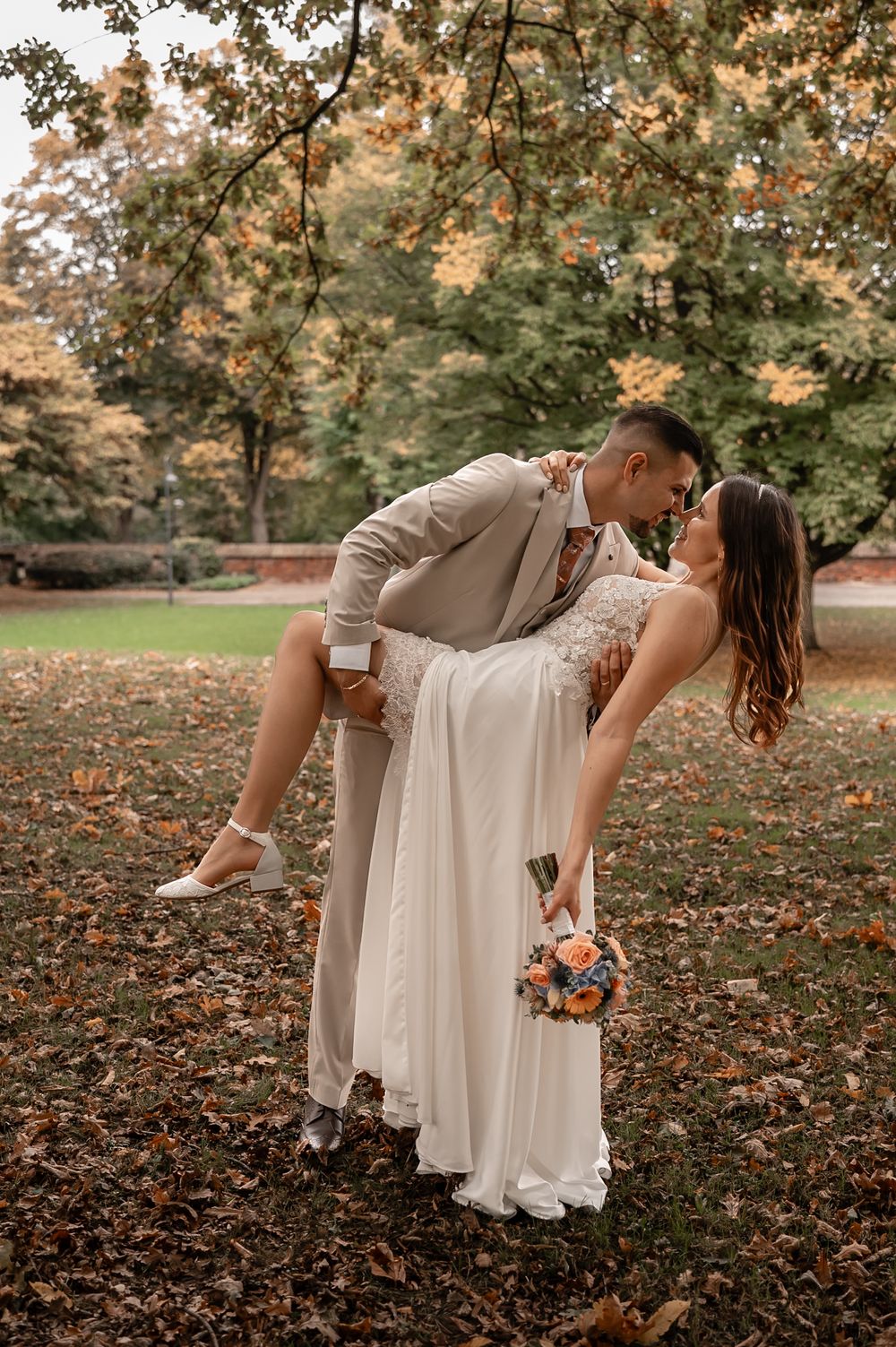 Newlyweds share a romantic dip pose surrounded by autumn foliage in a park setting.