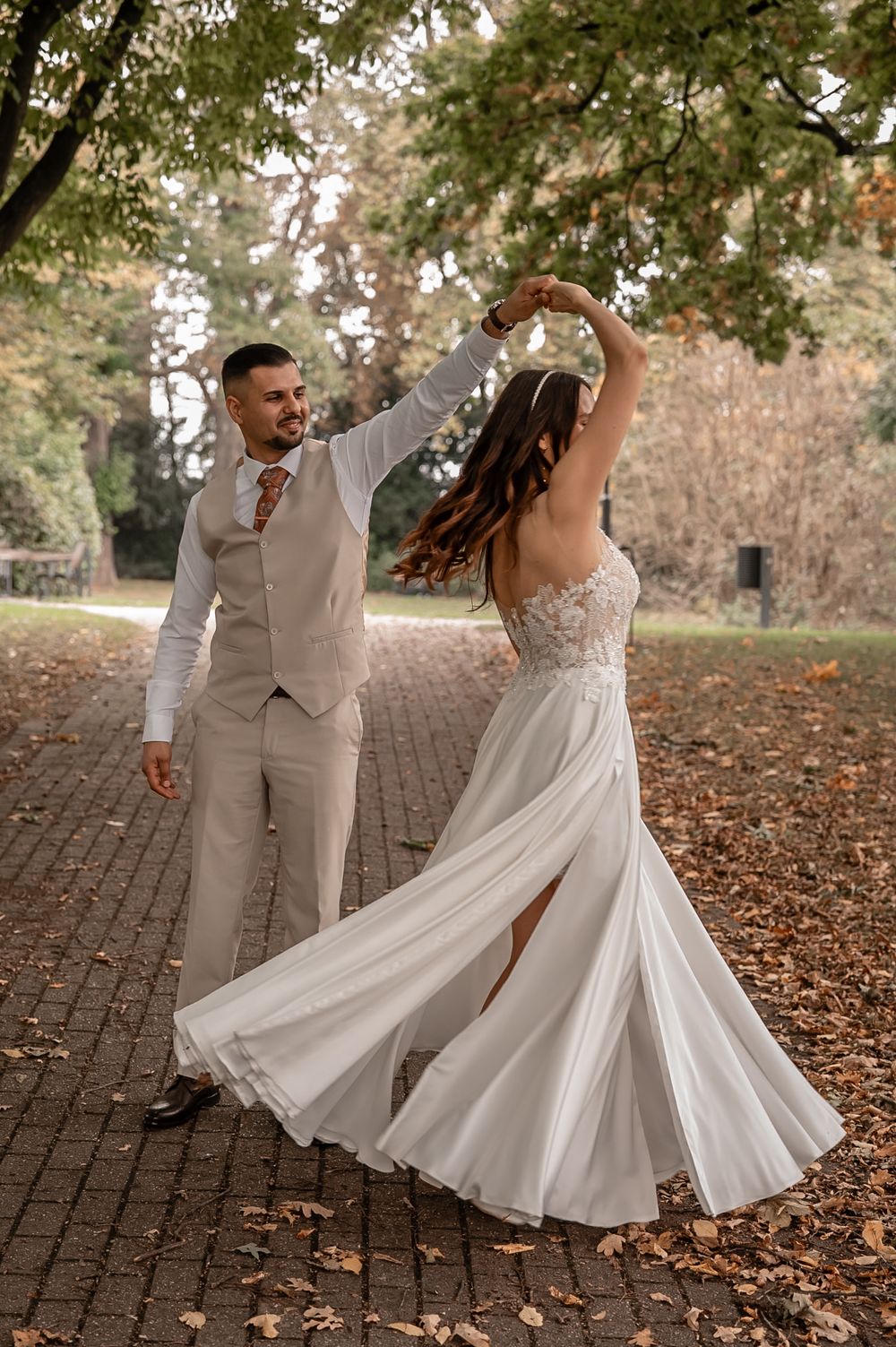 Newlyweds dance playfully among autumn leaves on a brick path with flowing white dress.