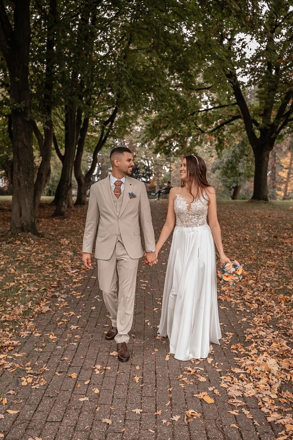 A couple walks hand in hand on a brick path through autumn trees, wearing elegant wedding attire in beige and white.