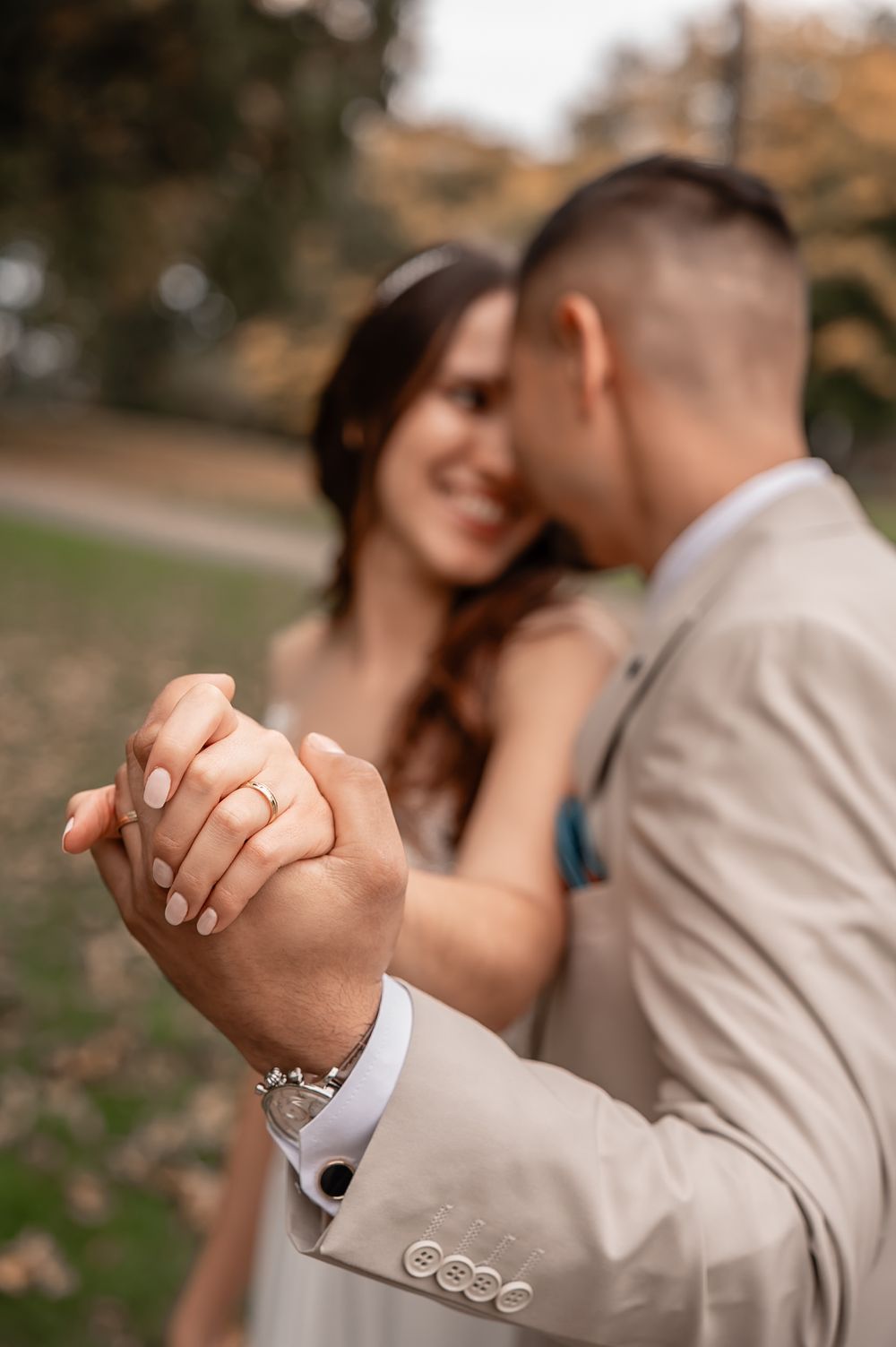 A couple holds hands intimately in an autumn setting with fallen leaves and soft bokeh background.