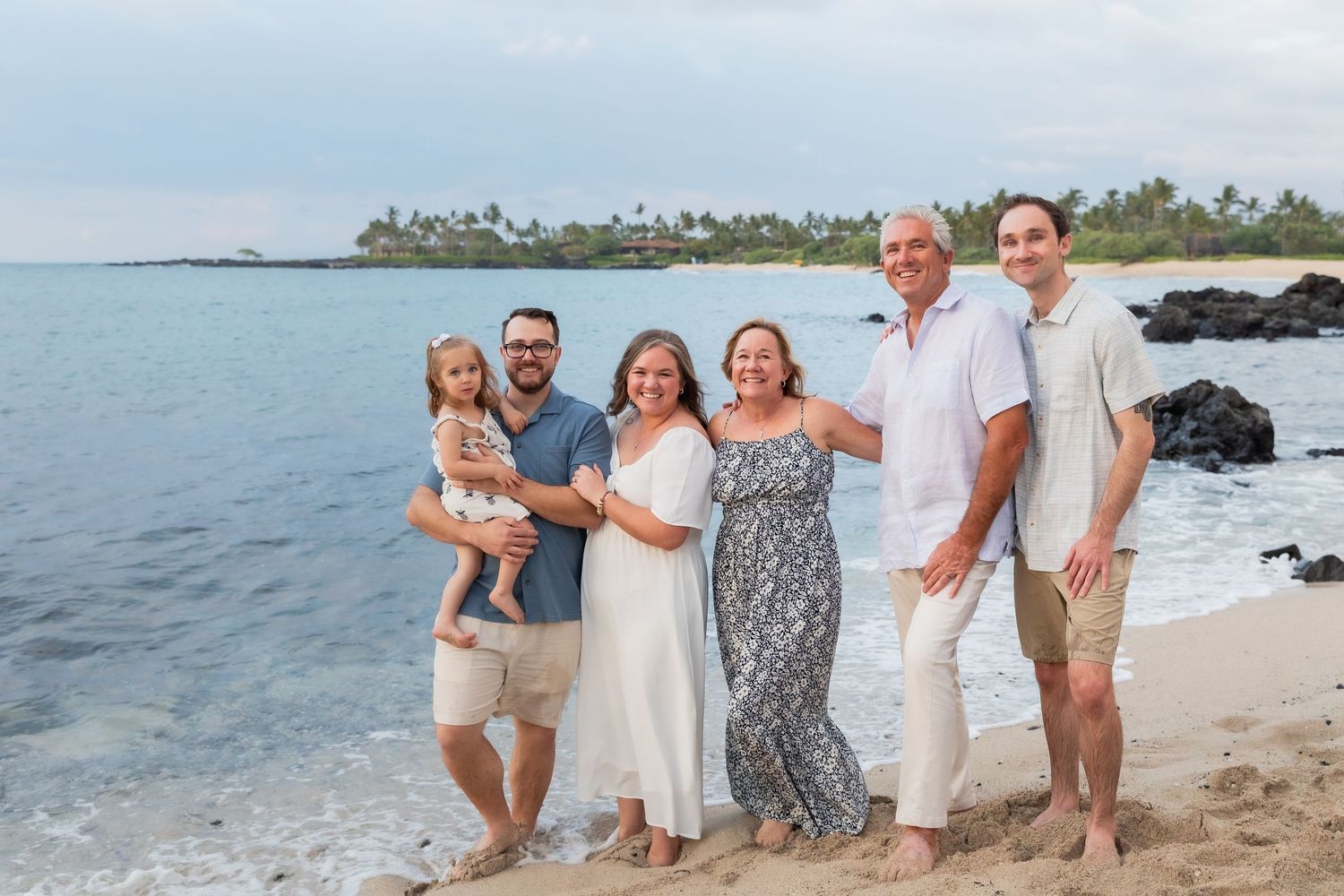 A group of friends wearing light floral and patterned dresses share joyful laughter together on a beach at sunset.