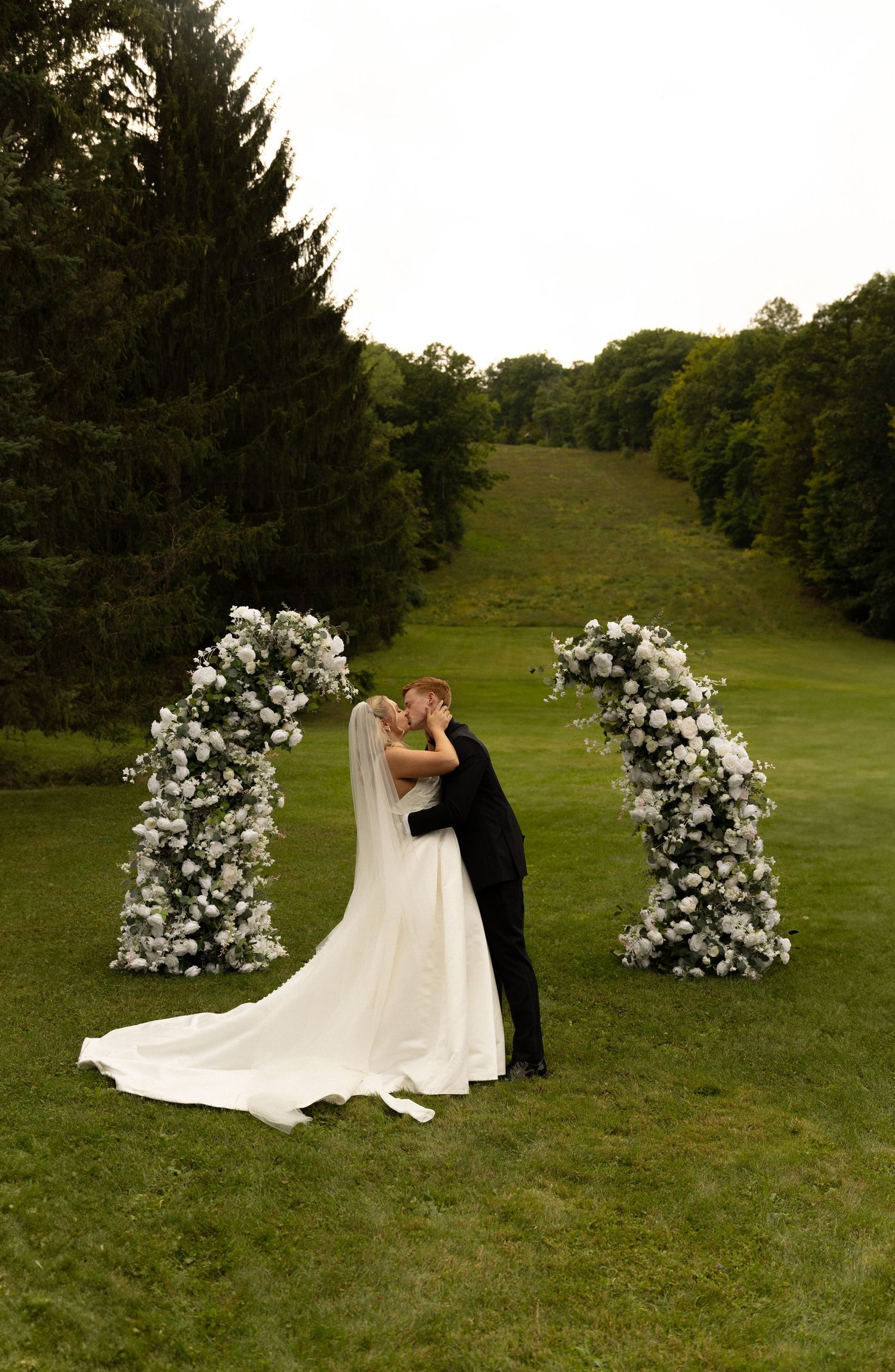 A romantic wedding scene with floral arches and a long white dress train flowing across green grass.