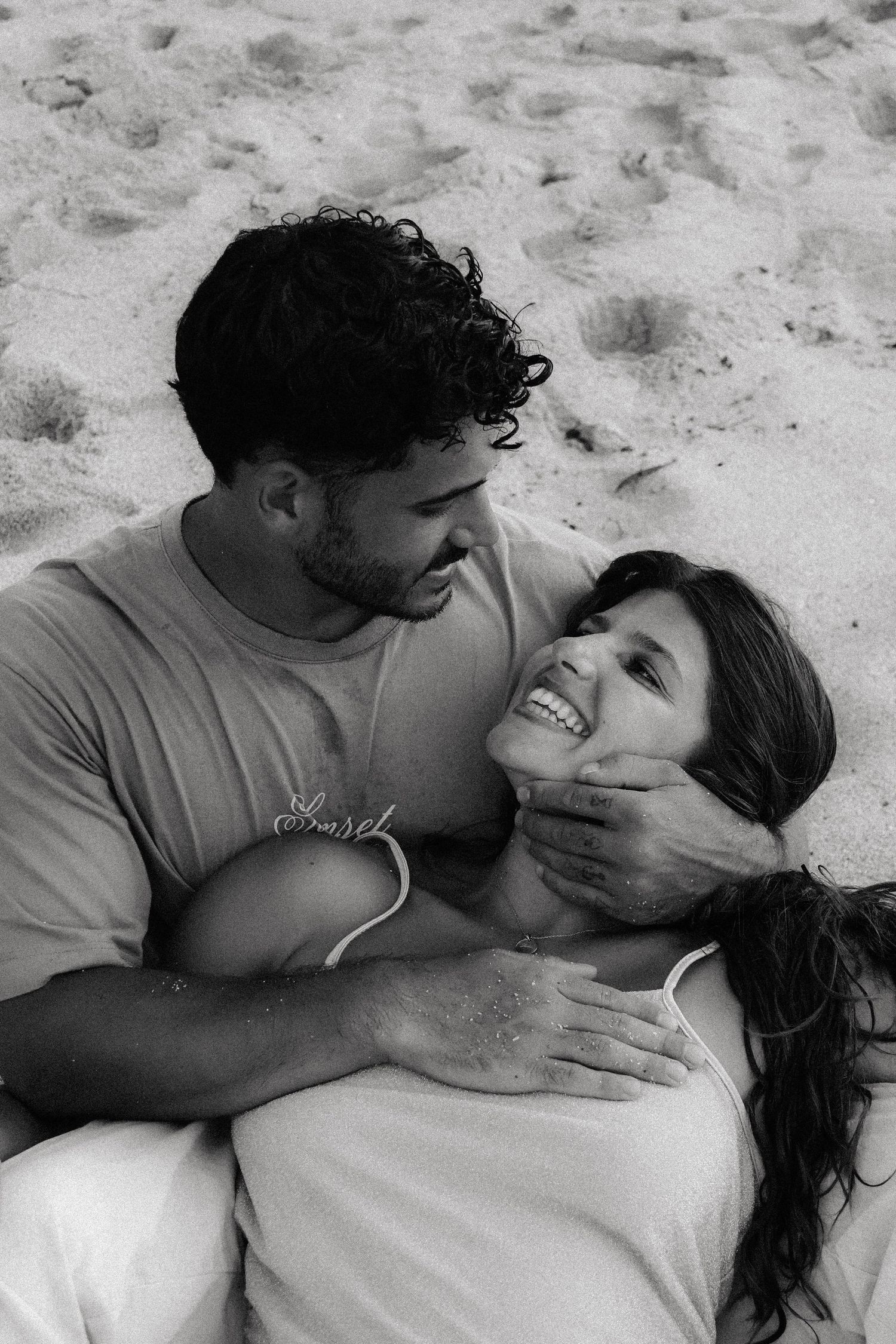 Black and white photo of couple sitting on beach sand, man gently holding womans face as she laughs.
