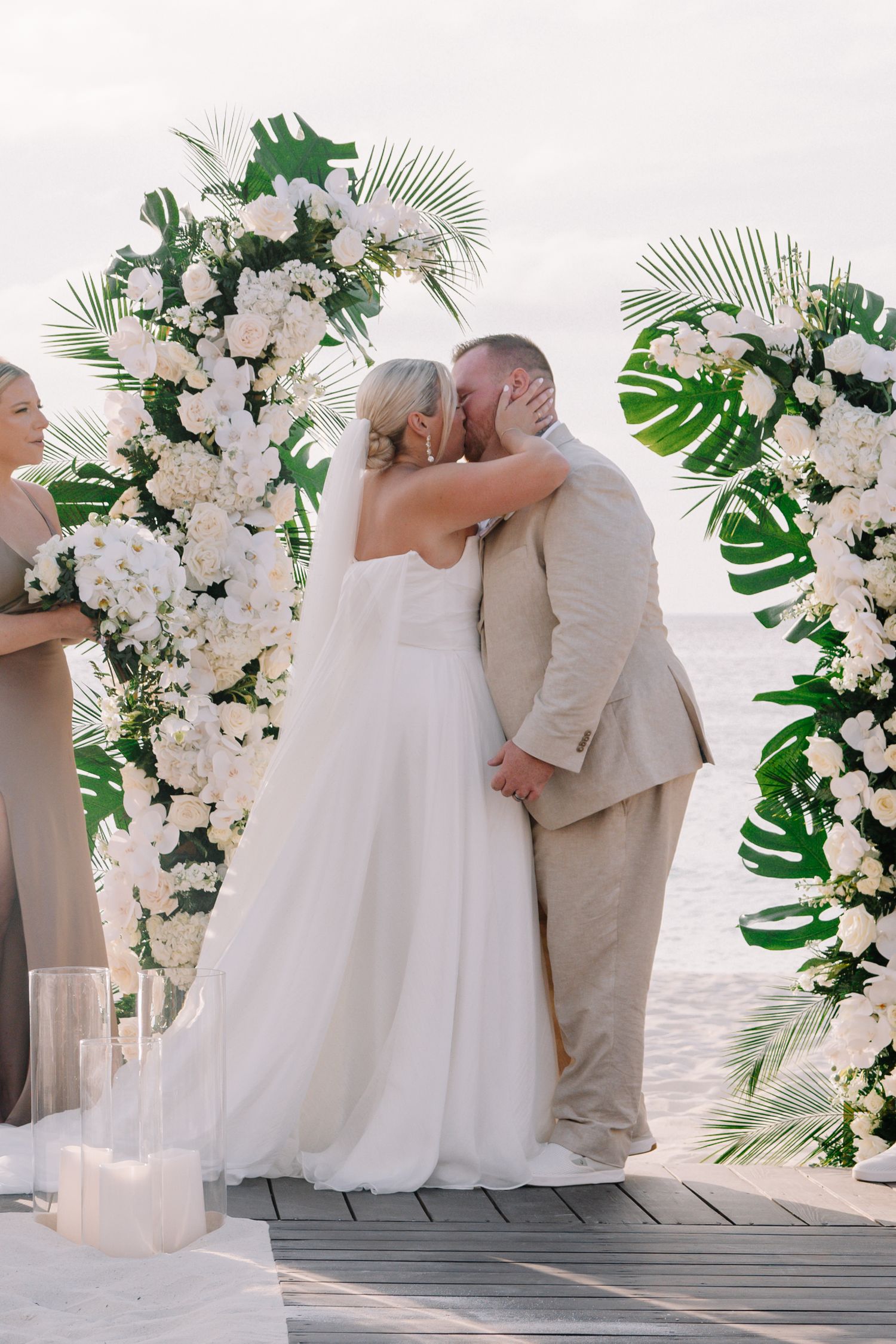 Wedding ceremony setup on beach boardwalk with white floral arch and tropical greenery decorations.