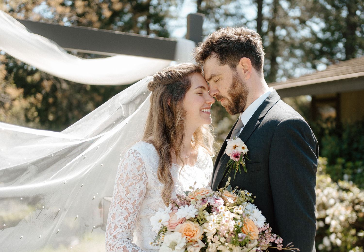 Bride and groom nearly kissing as a veil flows in the breeze outdoors.