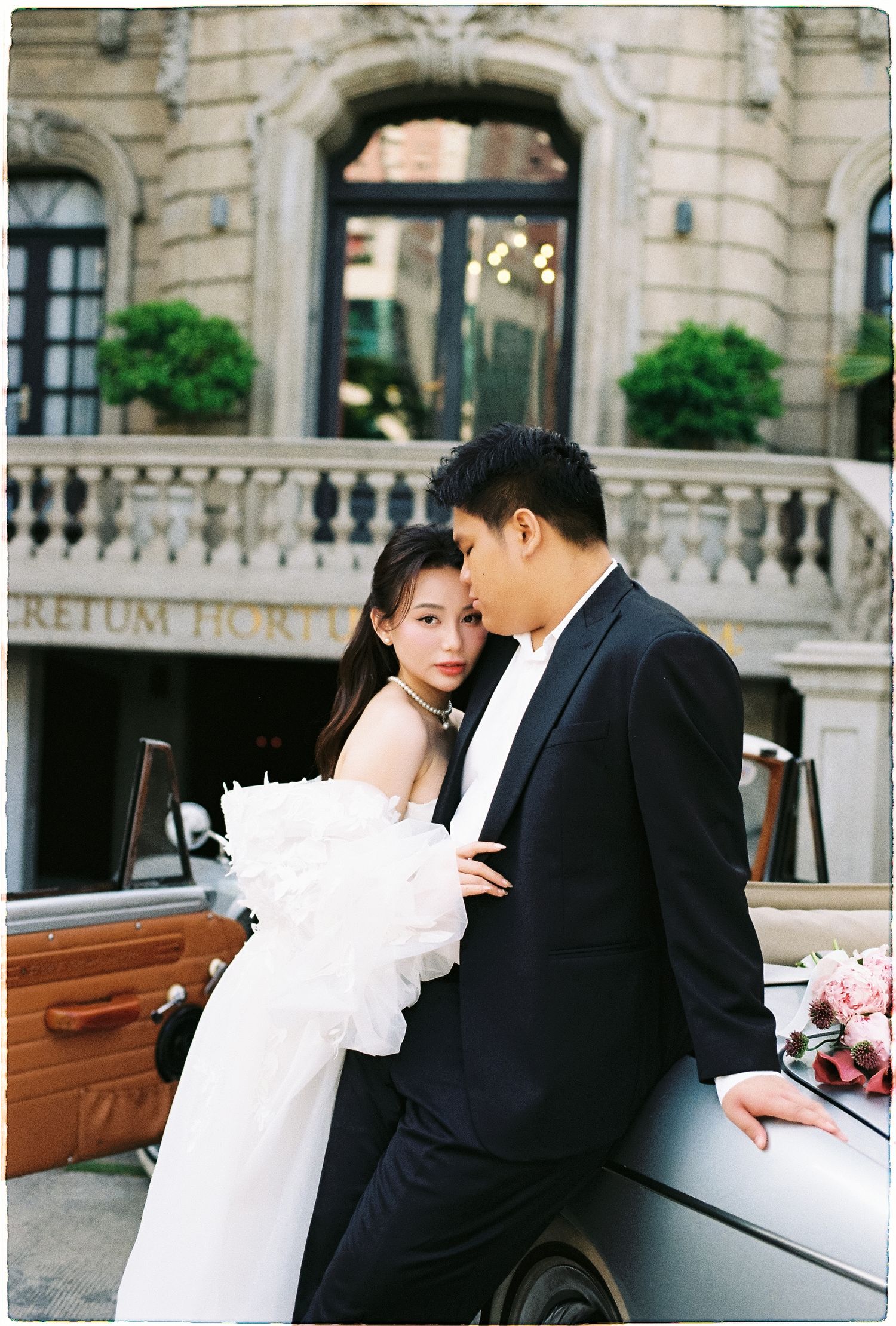 Bride in strapless white gown and groom in black suit posing beside vintage car at luxury hotel entrance.