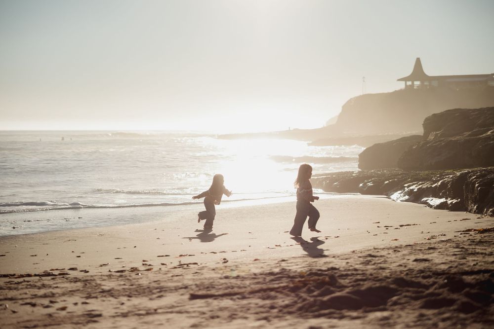 Silhouettes of people playing on a scenic beach at sunset with a lighthouse perched on coastal cliffs in the background.