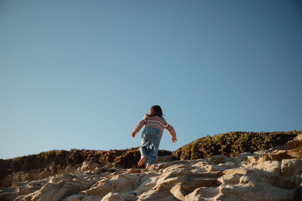 A person stands on a rocky cliff edge overlooking a scenic coastal landscape at sunset.