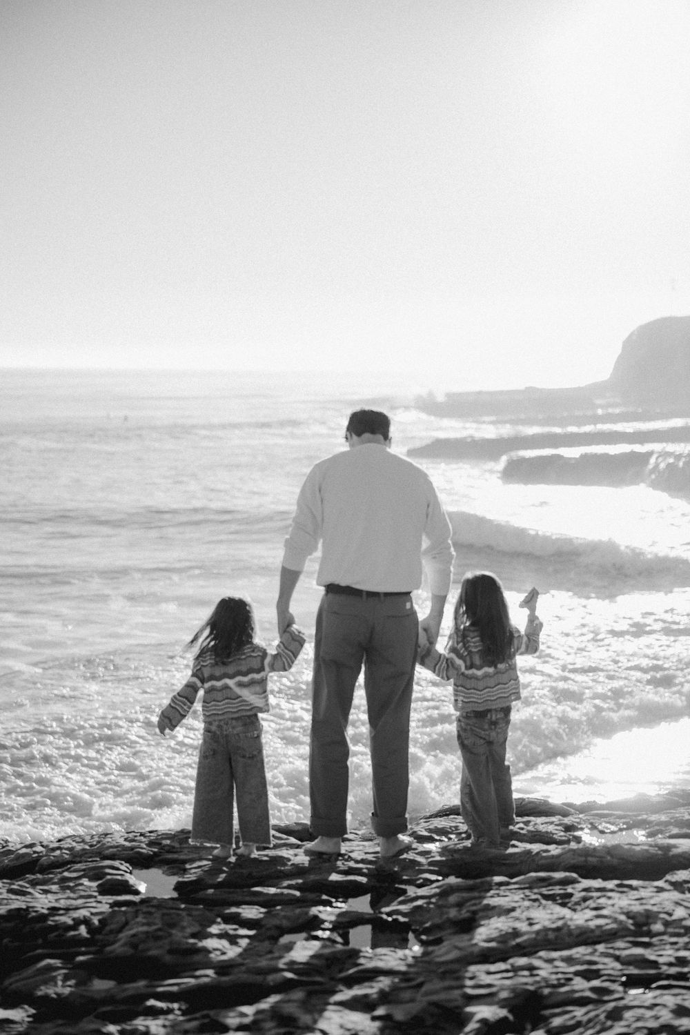 Black and white silhouettes of a family walking together along a rocky coastline at sunset.