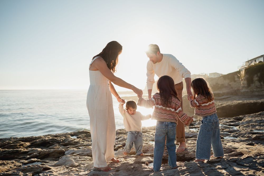 Family walking along rocky beach at sunset holding hands with ocean waves in background.