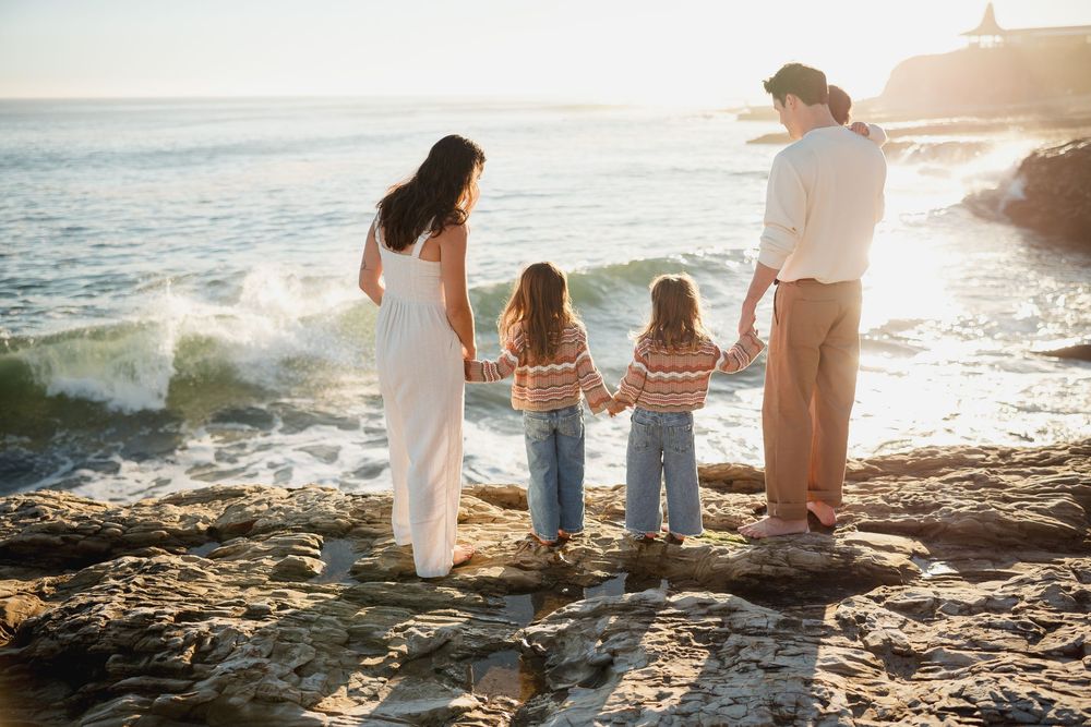 Family members sitting together on coastal rocks watching ocean waves at sunset.