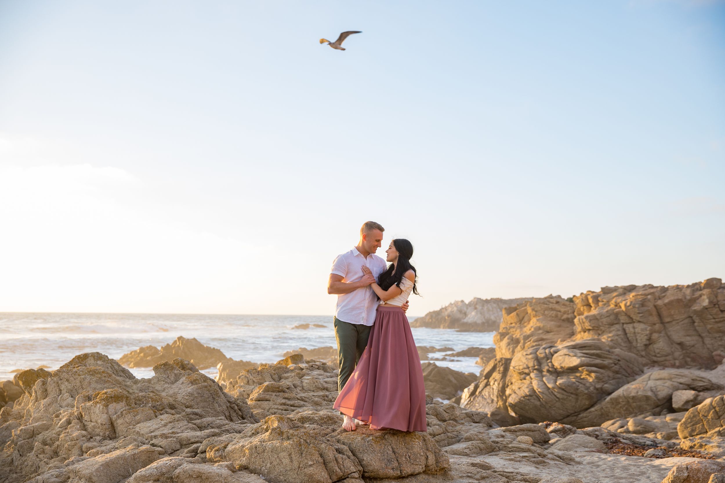 Couple embraces on rocky coastline at sunset while seagull flies overhead against blue sky and ocean backdrop in Pebble Beach, California.