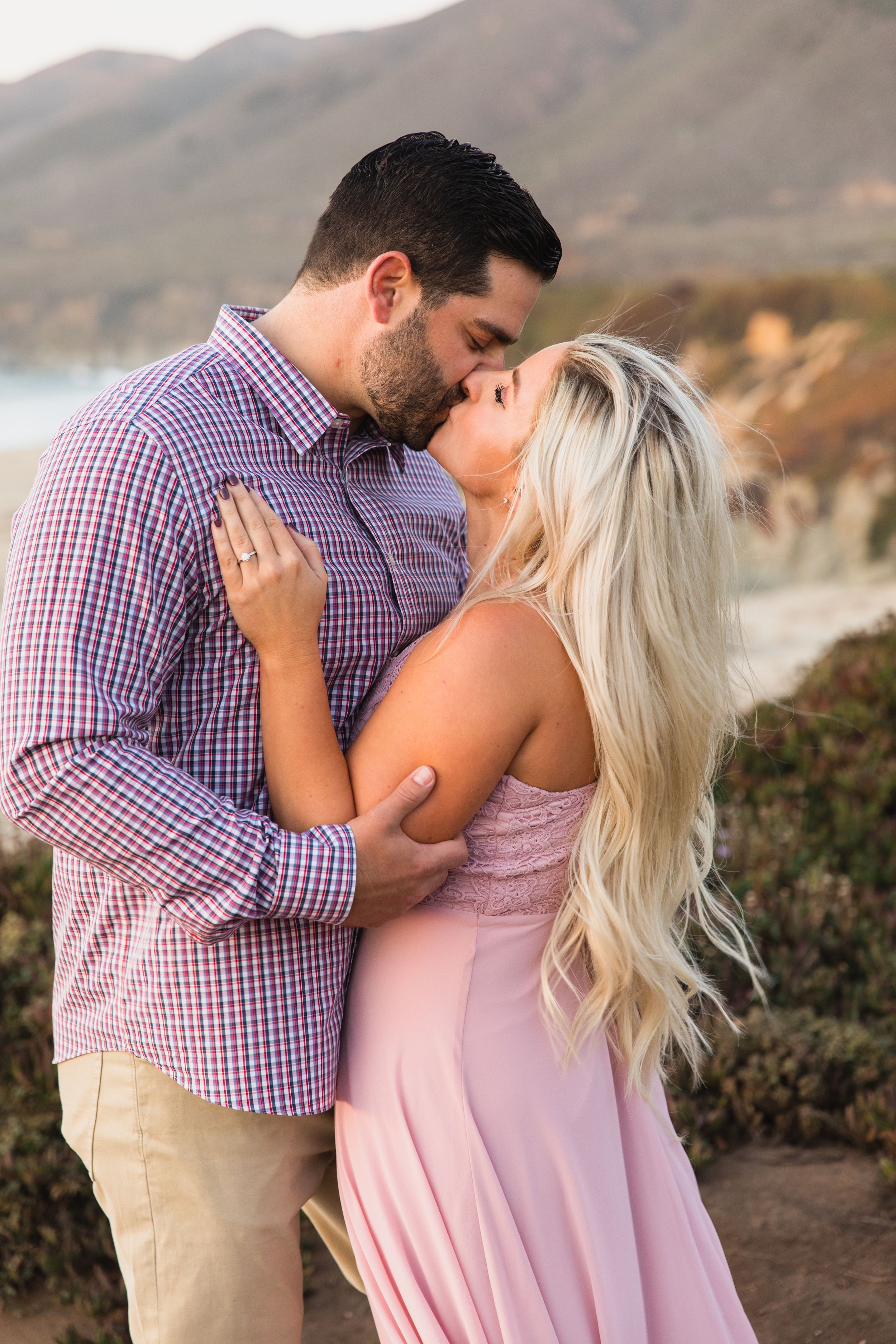 A couple shares a romantic embrace and kiss against a scenic mountain backdrop during sunset during a couples photo shoot.