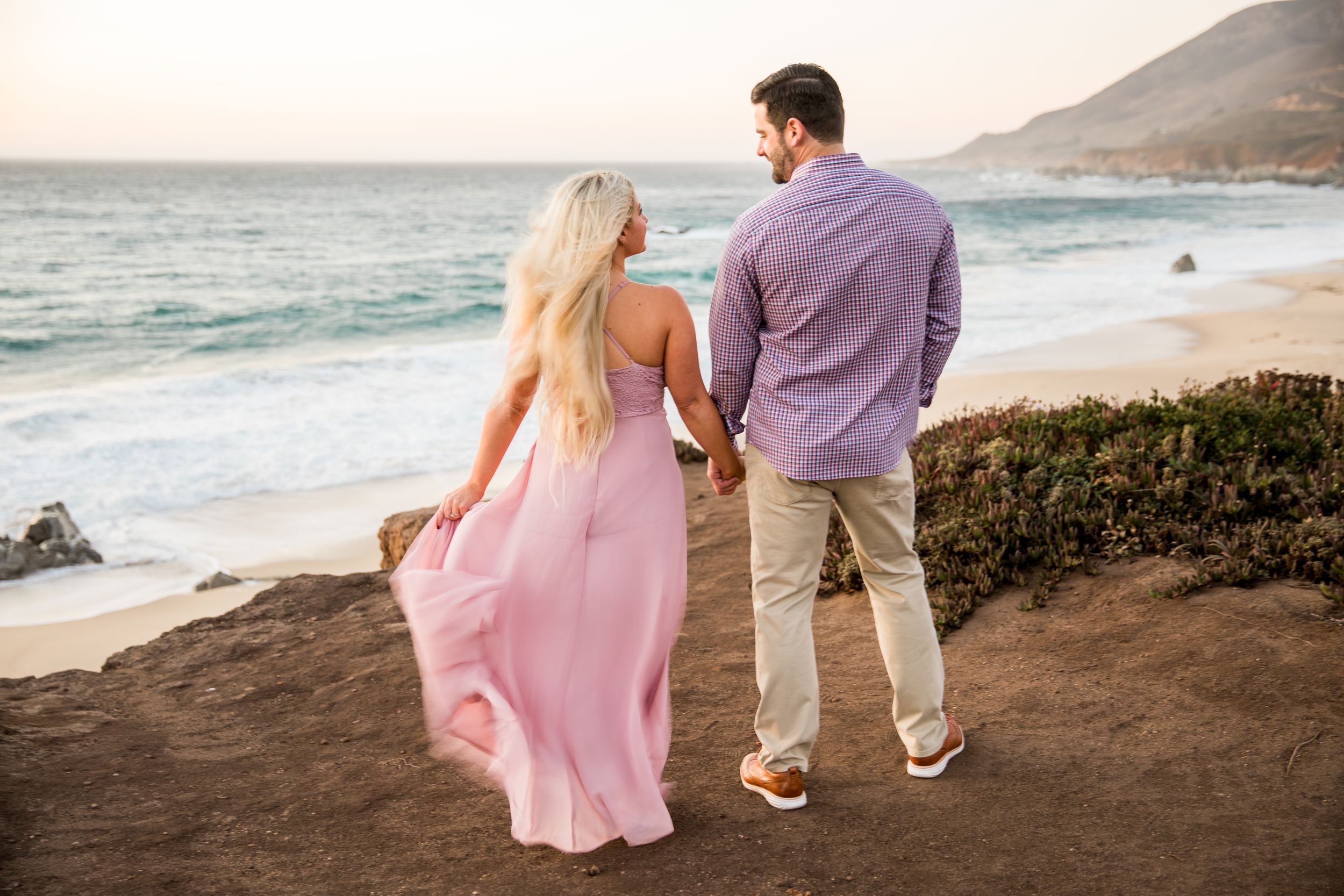 A couple stands on a coastal cliff overlooking the ocean at sunset as a pink dress flows in the breeze in Big Sur, California.
