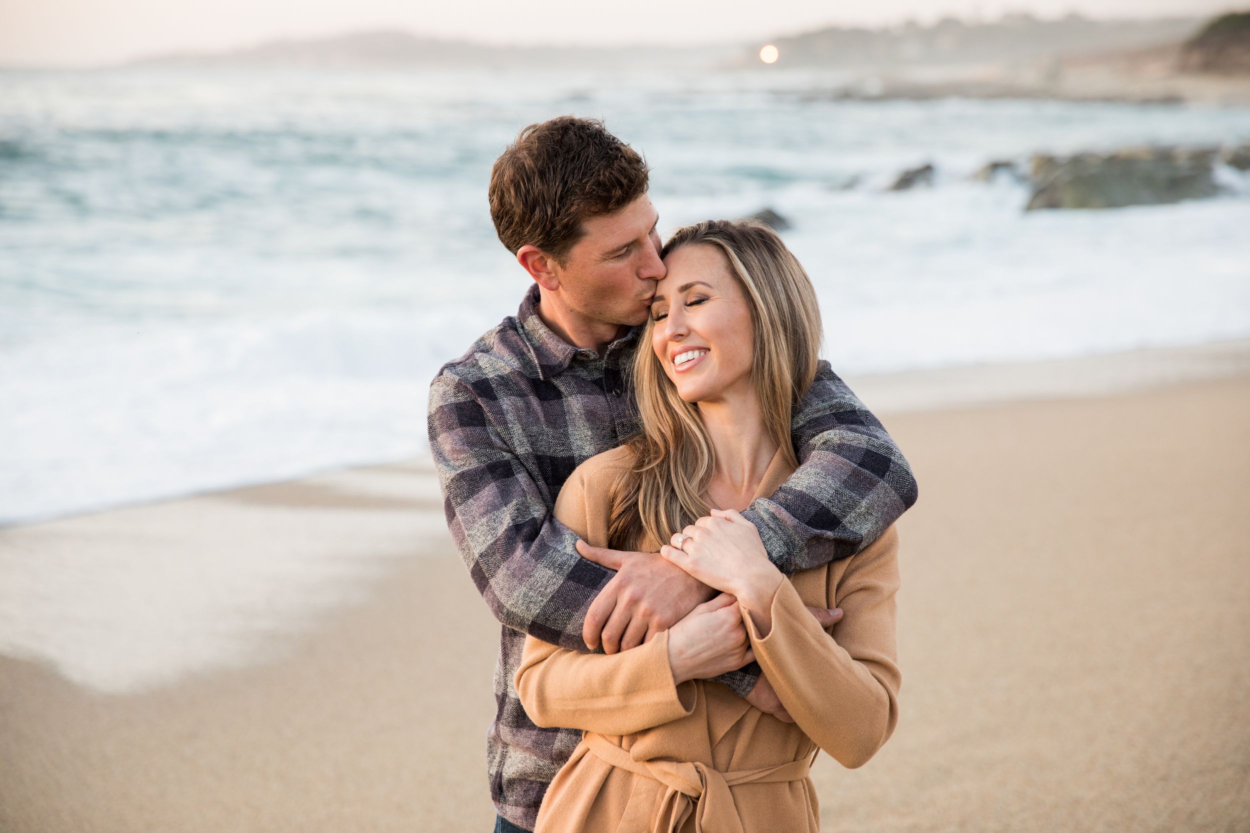 A couple embraces in a tender moment on a peaceful beach at sunset, with waves and misty coastline in the background at Carmel Beach.
