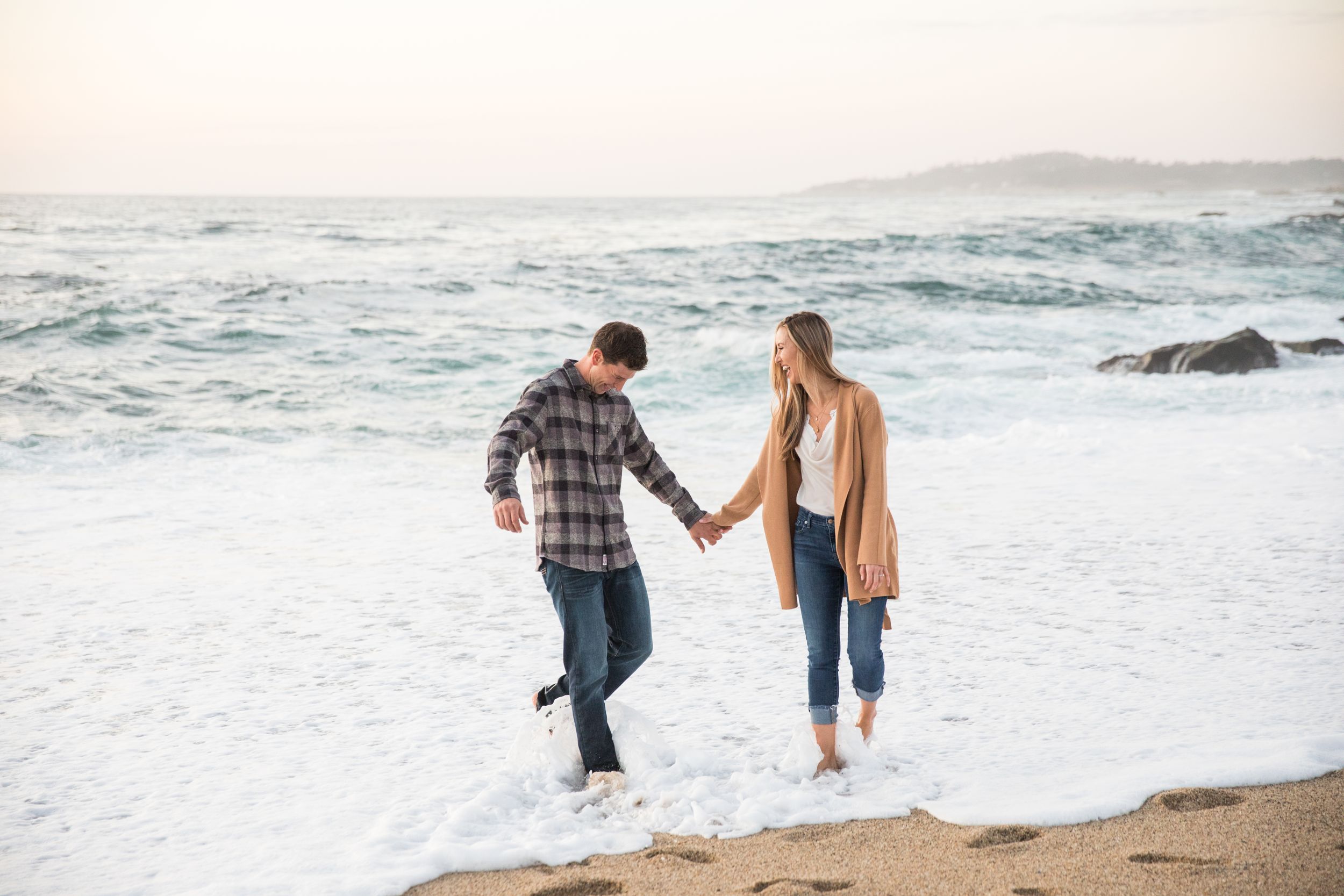 A couple holds hands while splashing in the ocean waves along a misty beach at sunset at Ribera Beach in Carmel.