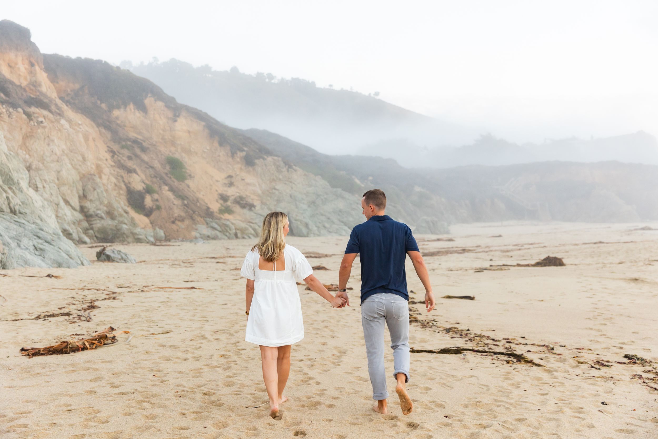 Two people holding hands while walking on a misty beach with coastal cliffs in the background for a couples shoot in Big Sur, CA.