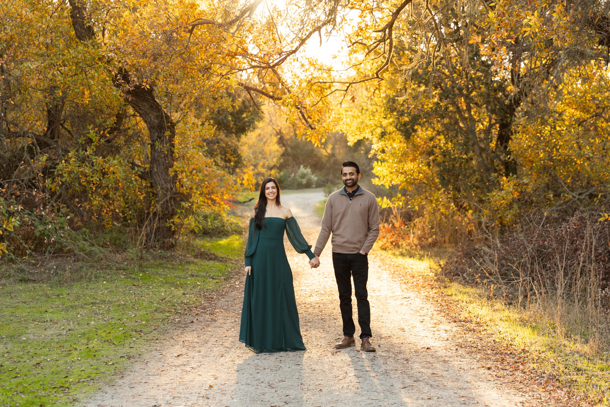 A couple holding hands stands on a sunlit path through autumn trees with golden foliage at sunset in Salinas, CA.