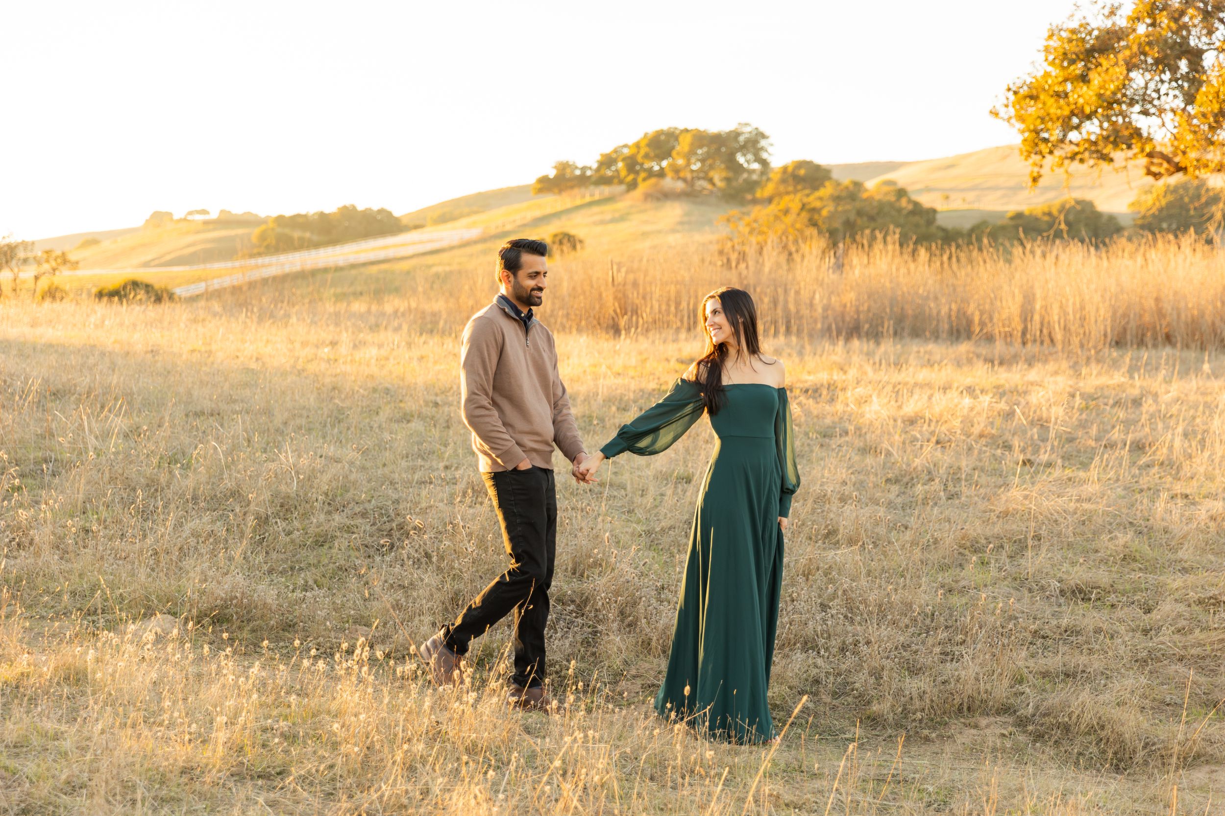 A couple shares a moment in a golden sunlit field during sunset, with the woman wearing an elegant emerald dress for couples photos.