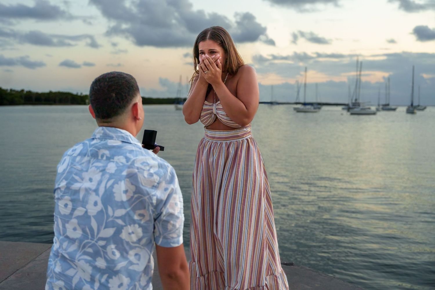 Man proposing by the water at sunset, capturing a surprised and emotional reaction from his partner.