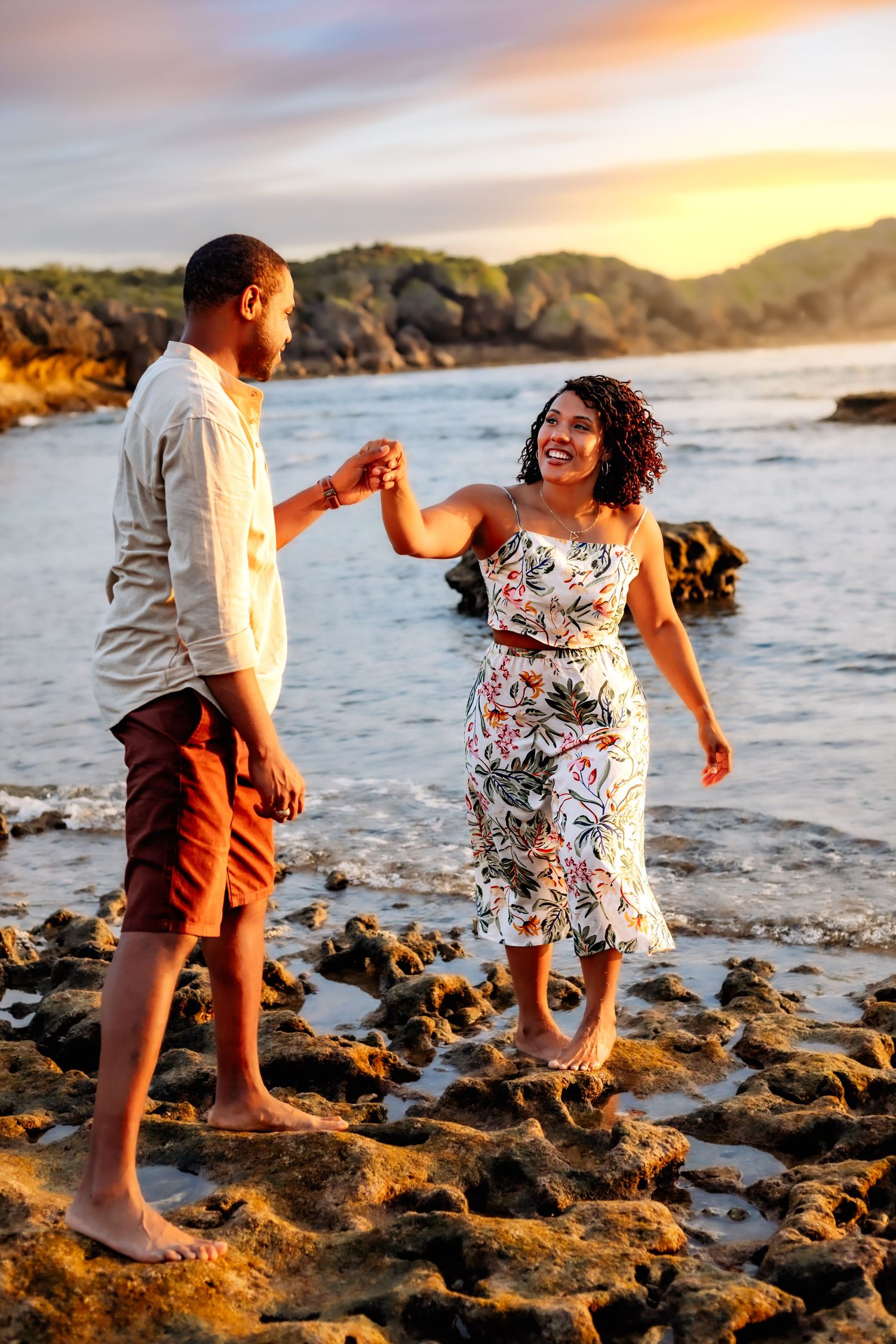 Couple dances and laughs together on a rocky beach during golden hour sunset.