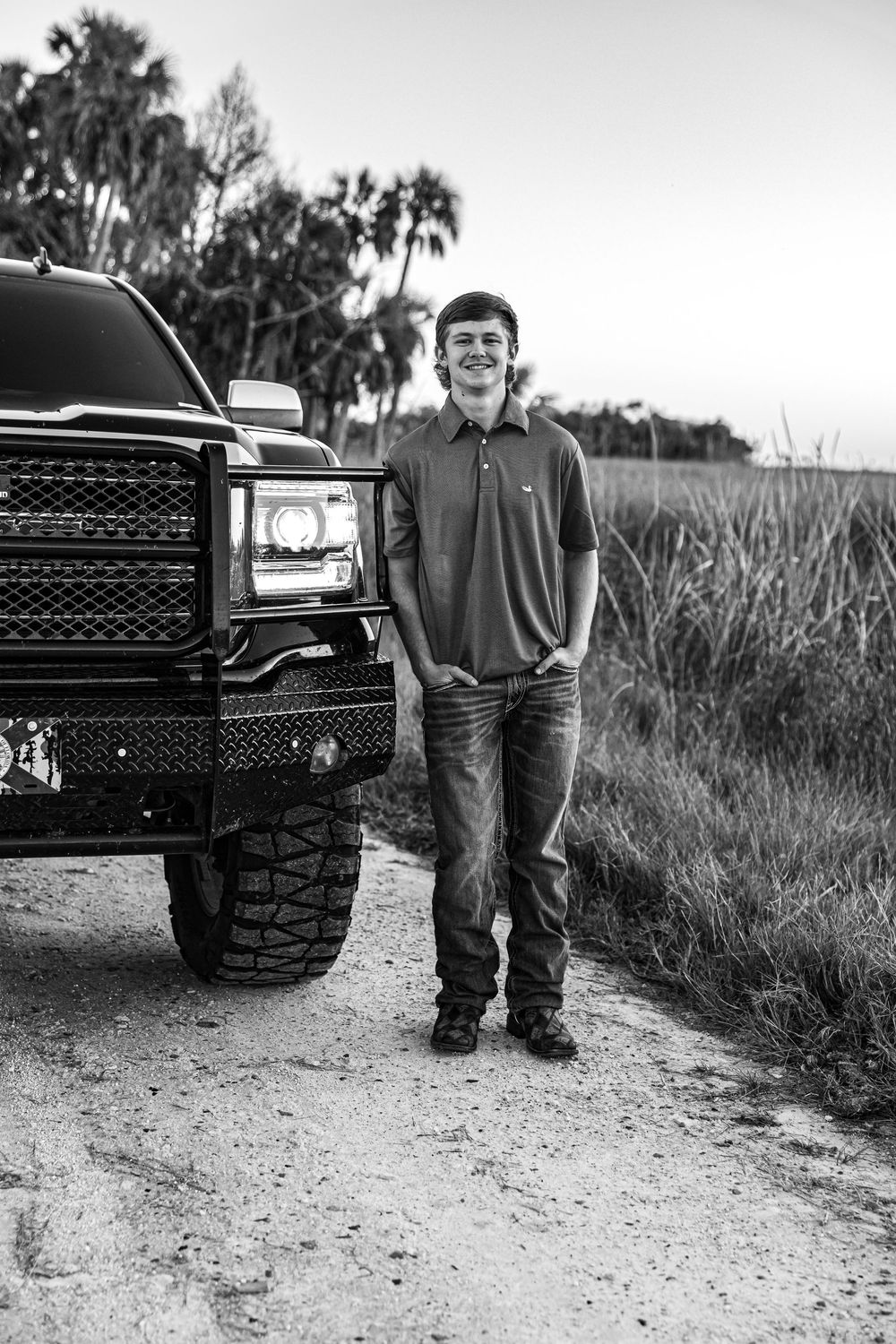 Young man smiling beside a lifted black truck on a dirt road in a black and white senior portrait.