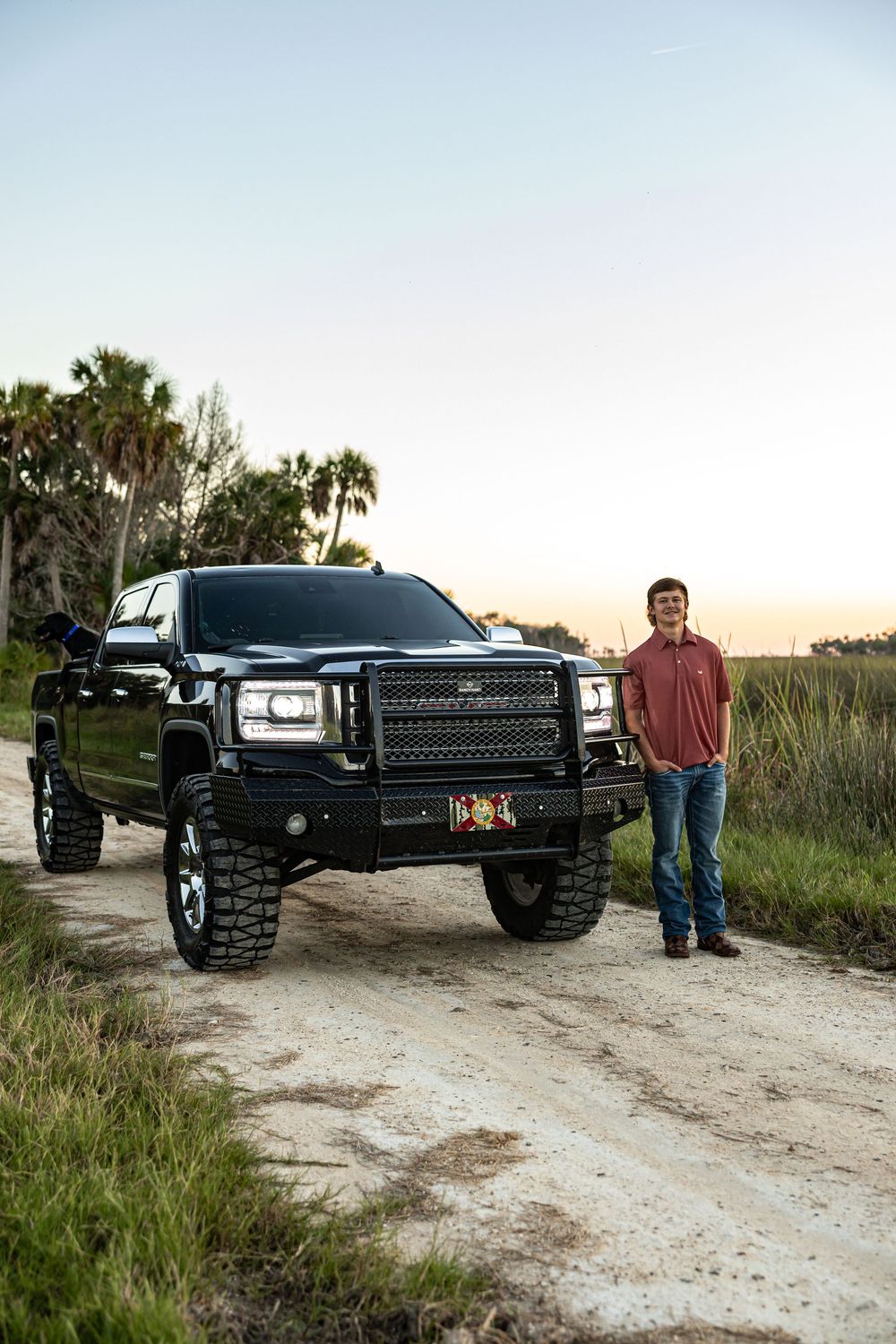 Young man in red polo stands beside a lifted black GMC truck on a rural dirt road at sunset.