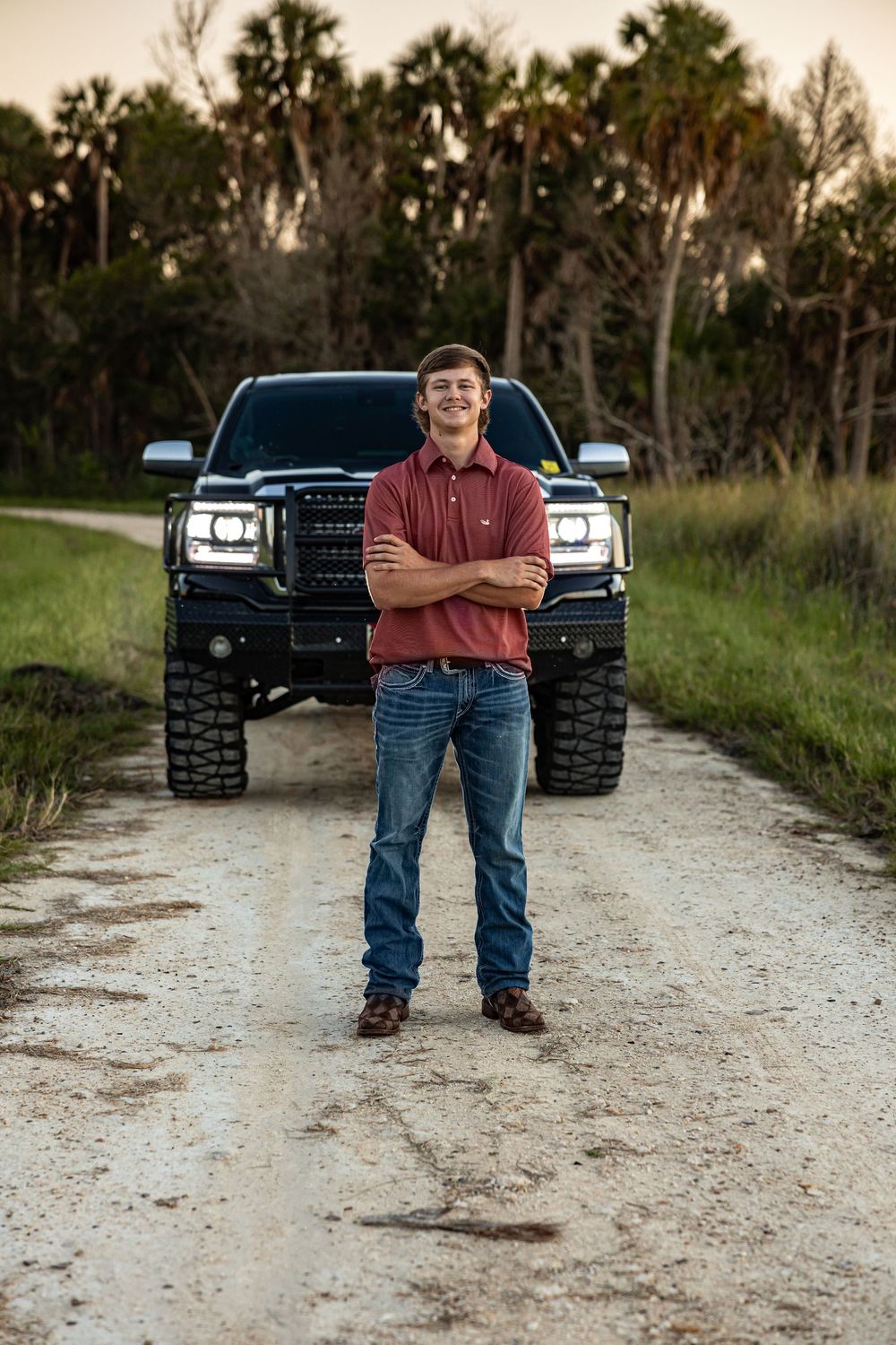 Smiling young man in red polo stands arms crossed before a lifted black truck on a dirt road in North Central Florida