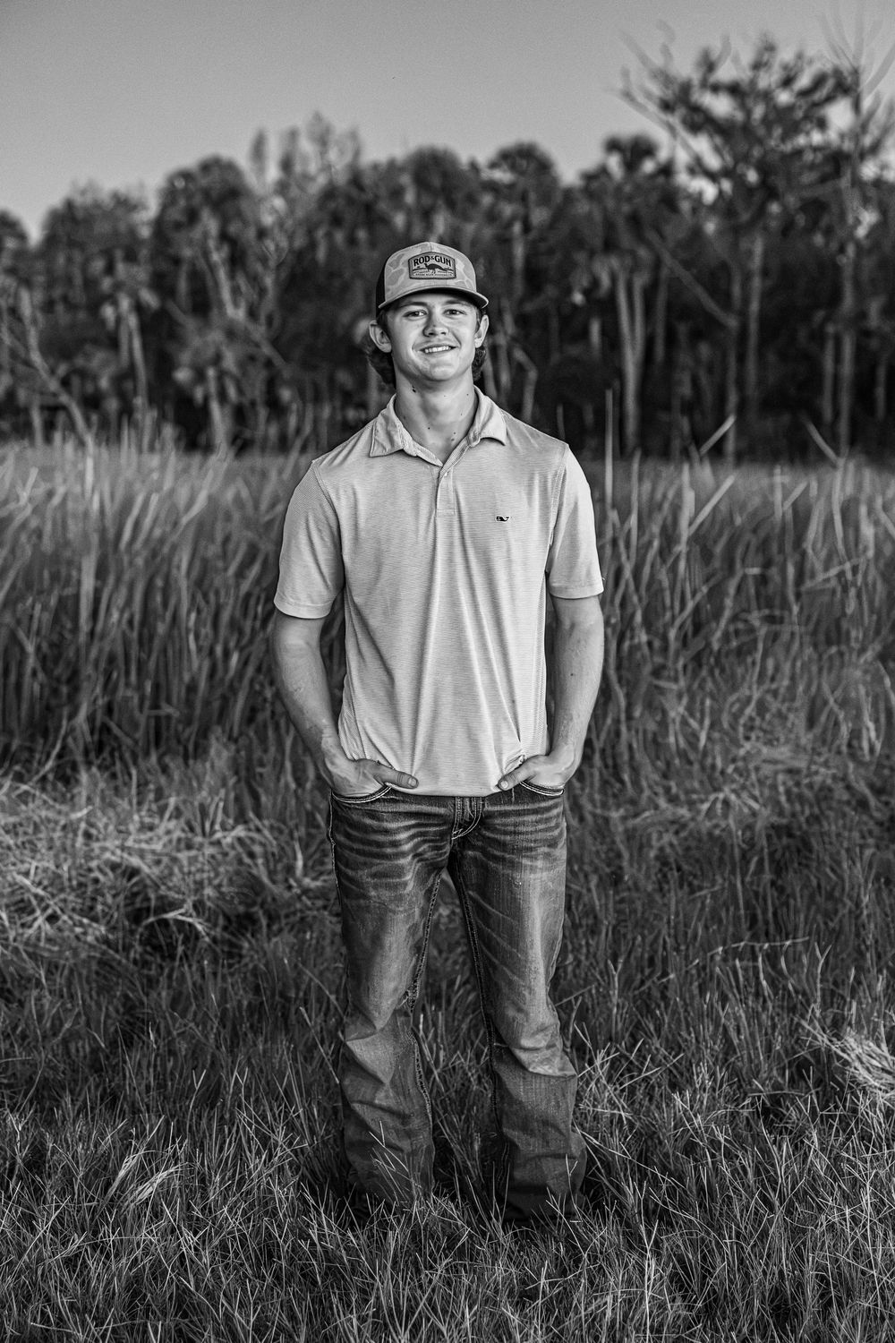 Young man in a cap and jeans standing in a grassy field in a black and white senior photo in Chiefland Florida.