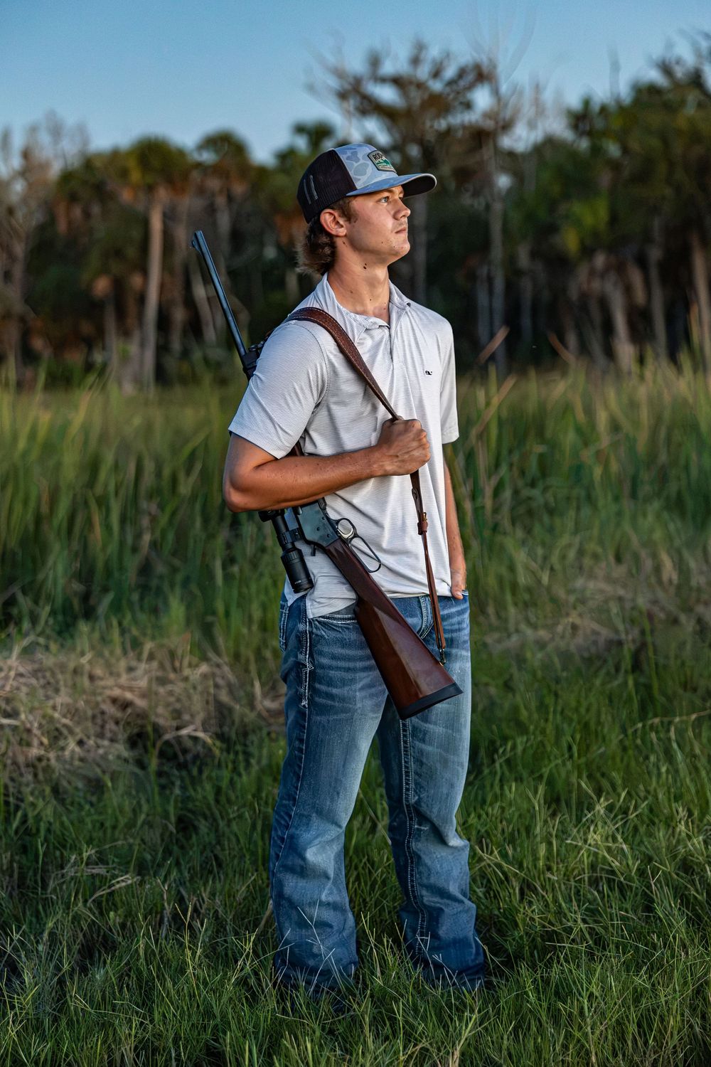 Chiefland High School Senior in jeans and cap holds a rifle over his shoulder standing in tall grass at golden hour in Chiefland, FL