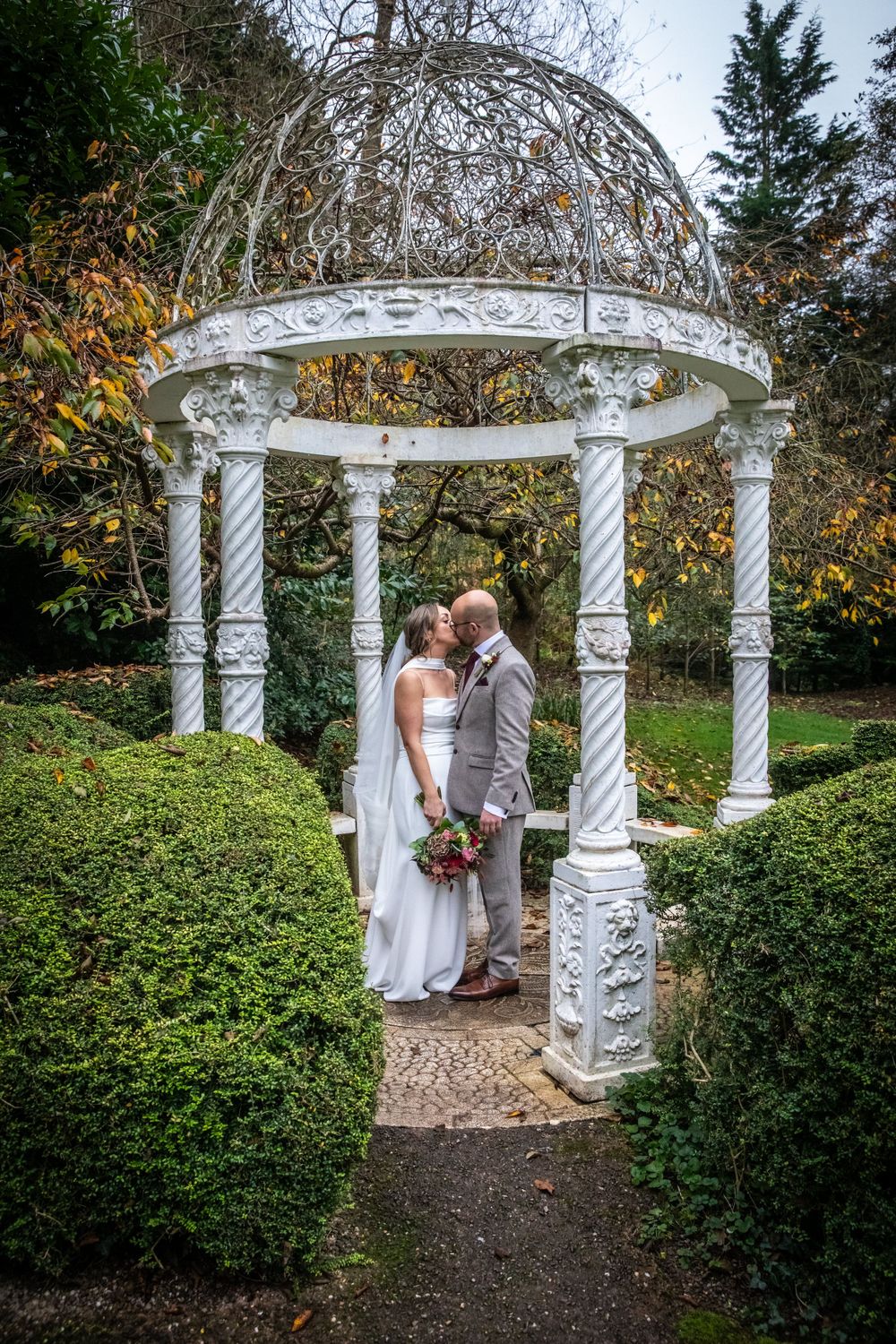 Couple shares a kiss under an ornate white gazebo surrounded by manicured hedges in autumn.